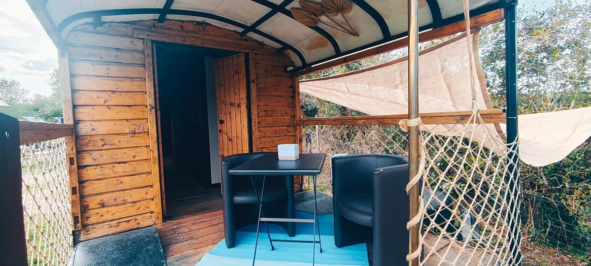 Le Luxury Circus — La terrasse couverte : table et fauteuils sous la voûte de bois, porte ouverte sur l'intérieur